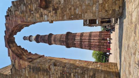 Towering Qutub Minar framed by a stone arch with visitors walking around its base.