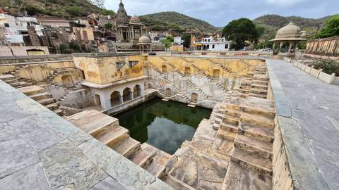 Historic stepwell and village buildings set against green hills under an overcast sky.