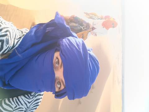 Close-up selfie of a woman in indigo desert scarf riding a camel across rolling sand dunes.