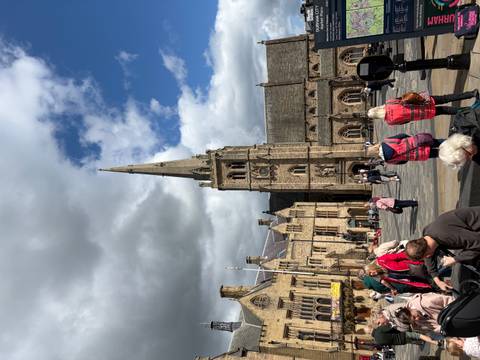 Busy town square with a tall church spire and clusters of tourists under mixed clouds