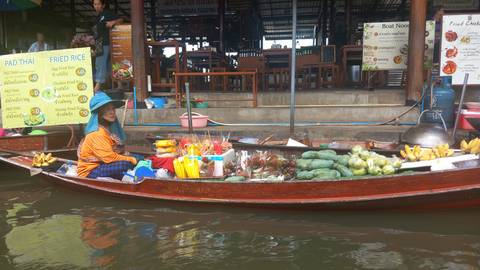 Vendor in orange shirt paddling a canoe laden with tropical fruit and snacks at a floating market.
