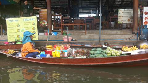 Vendor in a wooden boat selling fresh fruit and snacks at a floating market canal with restaurant stalls behind.