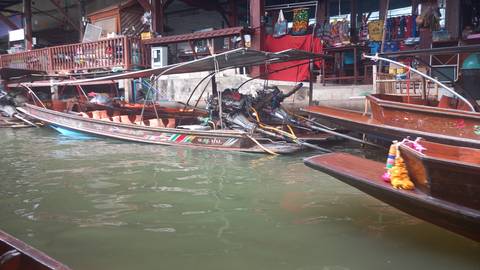 Long-tail wooden boats with exposed motors tied up along the pier of a floating market, green canal water in foreground.