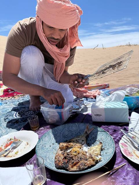 Close-up of a person preparing a beach picnic with grill rack, snacks and tea on a patterned mat in the sand.