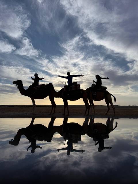 Silhouettes of three riders on camels at sunset, reflected in a shallow water pool beneath a dramatic sky.