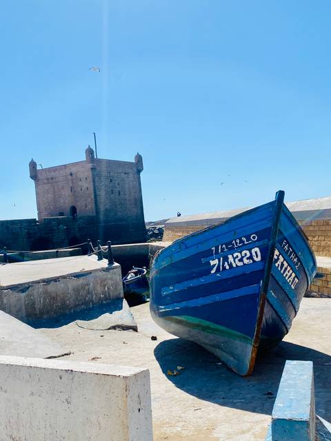 Blue wooden fishing boat moored beside the stone citadel tower of Essaouira under clear sky.
