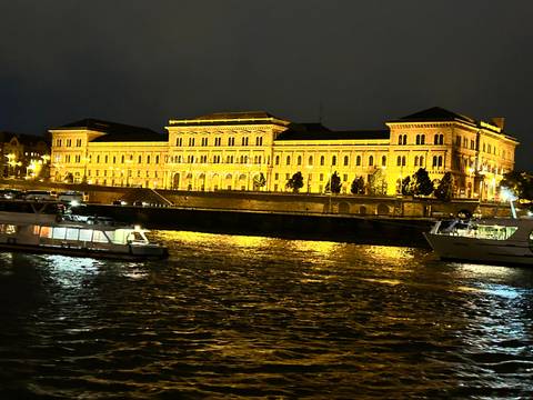 Illuminated riverside building in Budapest reflecting on the Danube at night with boats passing.