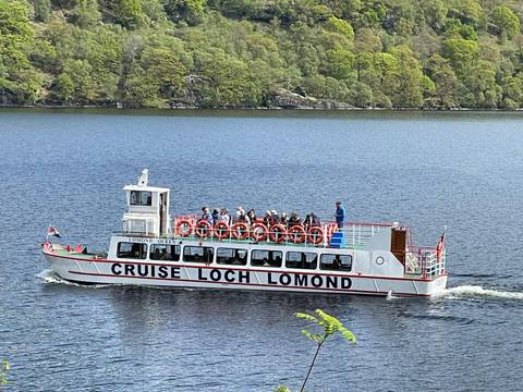Excursion boat 'Cruise Loch Lomond' carrying passengers across calm lake surrounded by forested shore.