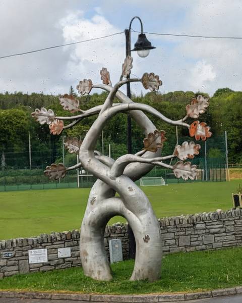 Metallic tree-like sculpture with entwined branches stands on a grassy sports field backed by forest.