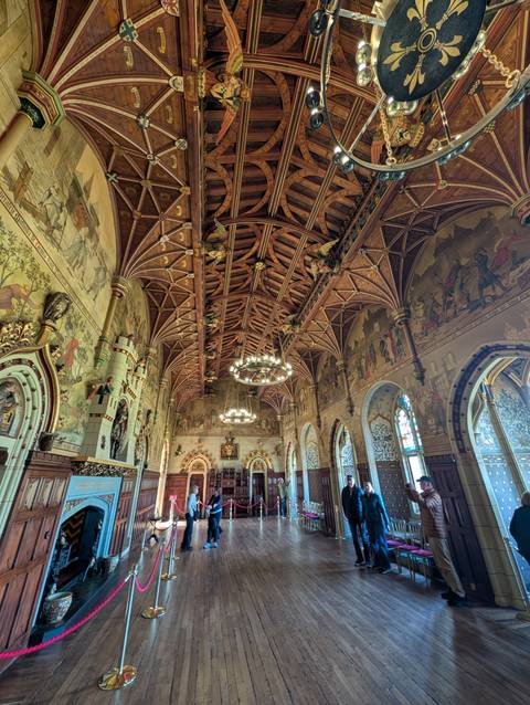 Opulent great hall with intricately carved wooden ceiling, chandeliers and painted murals.
