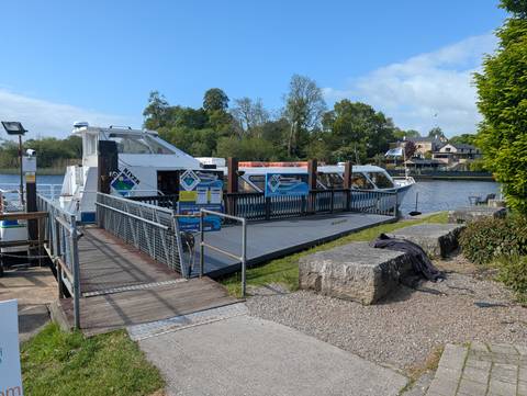 River dock with white tourist boats moored alongside a tree-lined bank under blue sky.