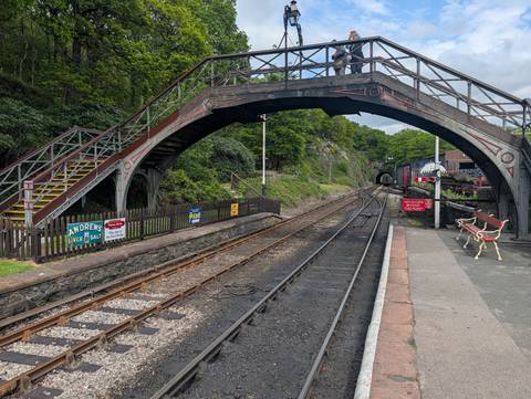 Vintage railway station with arched footbridge, tracks and forested rock cutting.