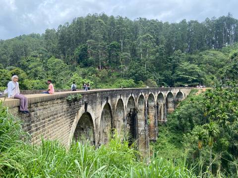 Historic nine-arch stone railway bridge spanning a deep forested gorge with visitors sitting along its edge