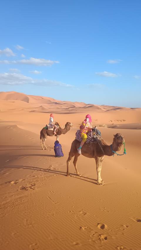 Two children riding camels led by a blue-robed handler amid sweeping orange dunes.