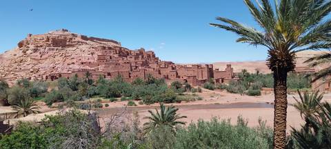 The mud-brick kasbah of Ait Benhaddou perched on a hill above a riverbed lined with palms.