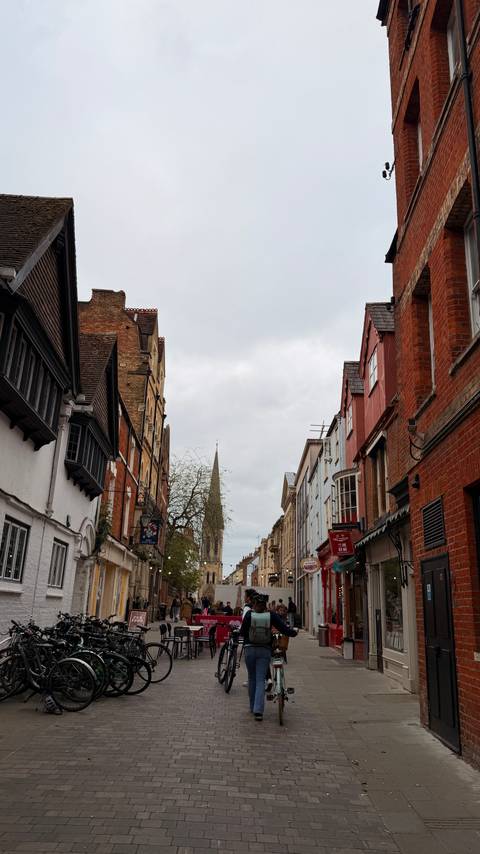 Historic brick and timber buildings line a narrow Oxford street that leads toward a distant church spire.