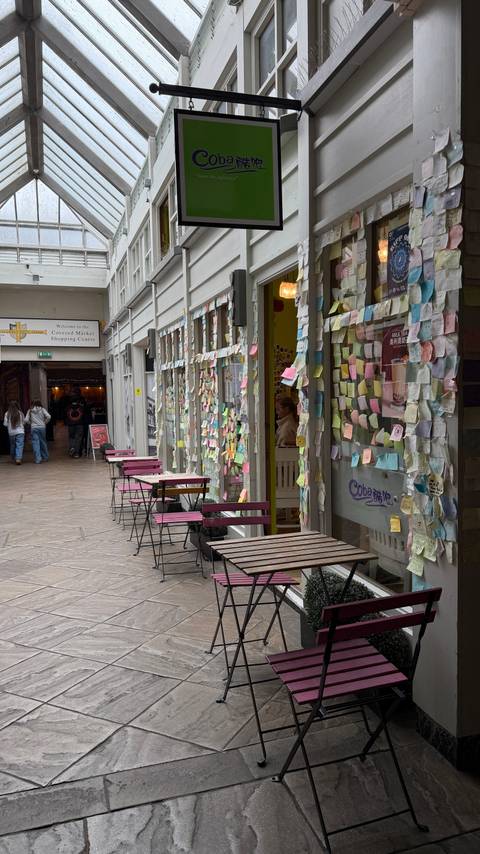 A cosy arcade where café tables face a shopfront plastered with colourful sticky notes and messages.