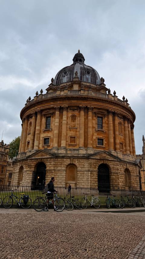 The iconic circular Radcliffe Camera stands in warm golden stone beneath a moody grey sky.