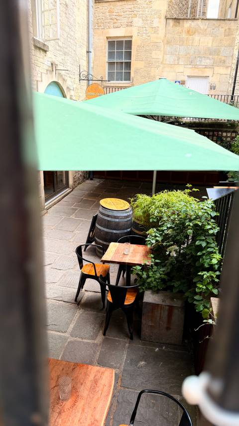 A small courtyard café with wooden barrel tables, greenery and a shaded umbrella-covered setting.