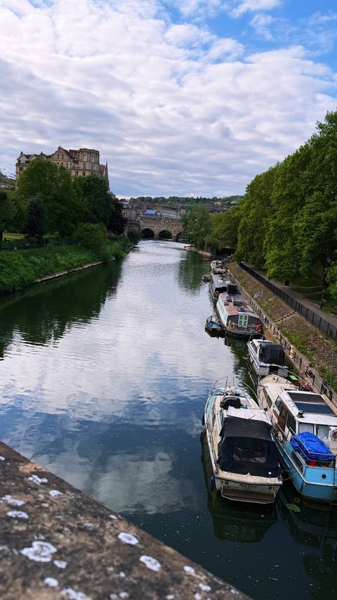 Calm river with moored narrowboats leads toward an arched stone bridge and historic Bath skyline.