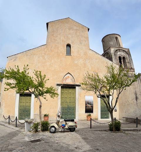 Simple stucco church façade with green wooden doors and young trees against a pale sky.