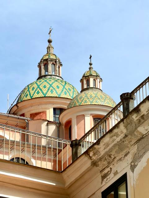 Colorful tiled domes of a Mediterranean church rise above peach-colored walls and railings against a clear blue sky.