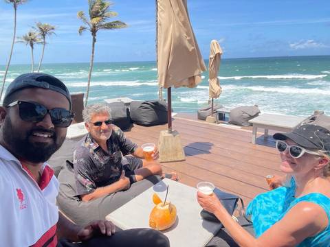 Three friends enjoy cold drinks on a beachfront deck overlooking waves and palm trees.