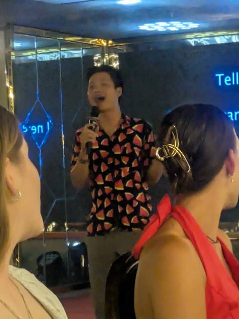 Indoor scene of a host singing into a microphone wearing a watermelon-print shirt during evening entertainment.