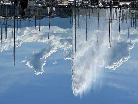 Geneva's Jet d’Eau fountain shooting water high beside sailboat masts with alpine backdrop and scattered clouds.