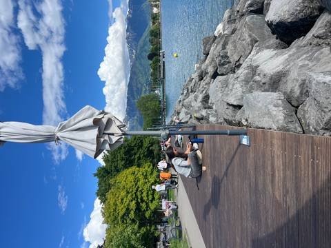 People relaxing and sketching on a wooden lakeside promenade with rocky shore and mountains beyond.
