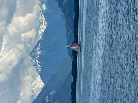 Single sailboat gliding across a blue alpine lake under towering snow-capped mountains and dramatic clouds.