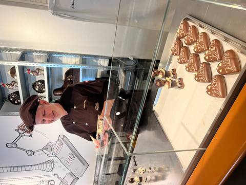 Smiling chocolatier crafting heart-shaped chocolate figures behind a glass counter that reads 'Je t’aime'.