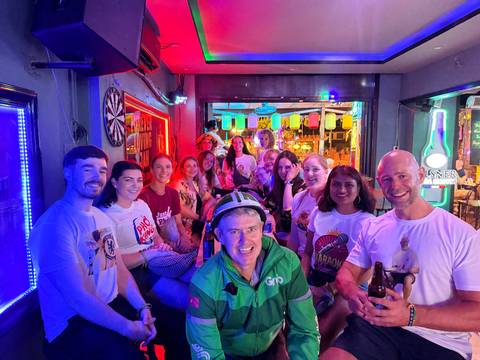 Tour group posing inside a neon-lit bar with bright decorative lights and drinks in hand.
