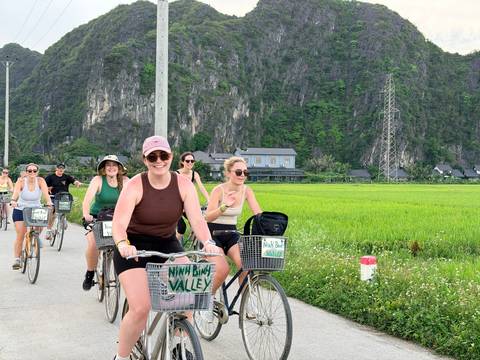 Cyclists riding past green rice fields and karst cliffs with 'Ninh Binh Valley' signs on their bikes.