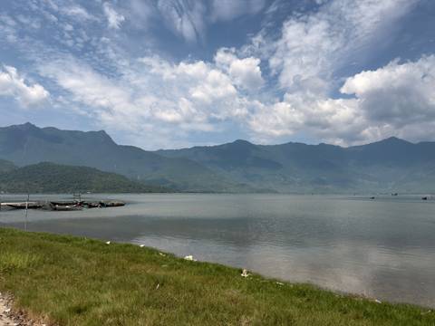 Calm lagoon lined by fishing boats with misty mountains rising under a partly cloudy sky.
