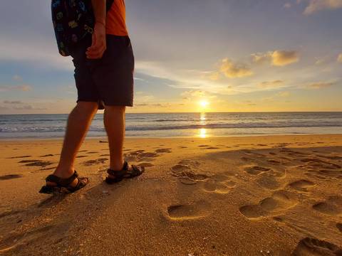 Close-up of a traveller's legs walking along a golden beach during a vivid sunset over the ocean.