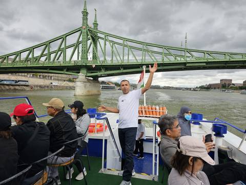 Passengers seated on an open-air river boat as it passes beneath a large green steel bridge on a cloudy day.