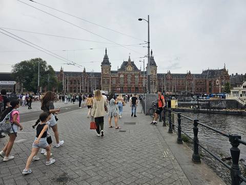Busy waterfront scene with people walking in front of a historic red-brick railway station beside a canal.