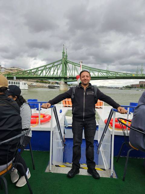 Man posing on the deck of a sightseeing boat with a green steel bridge and city skyline behind him.