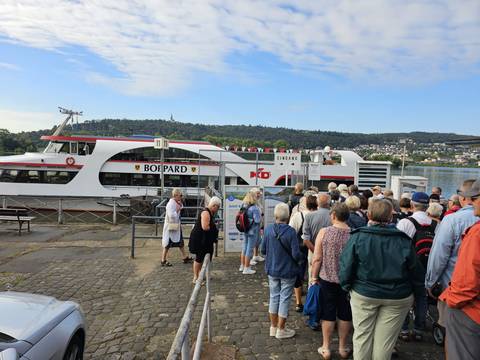 Line of tourists waiting to board a white excursion boat docked on a wide river.