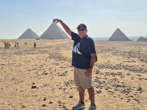 Tourist playfully posing to 'pinch' a pyramid at the Giza Plateau with multiple pyramids in view.