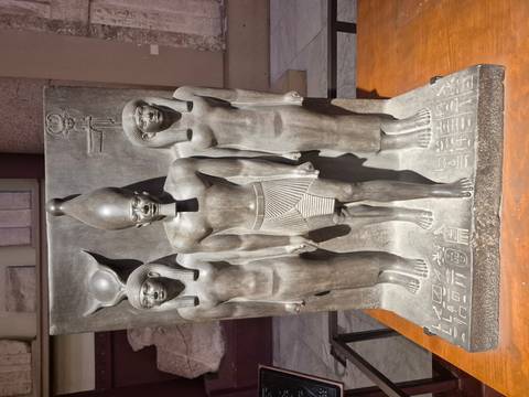 Dark stone triad statue of Pharaoh Menkaure flanked by two goddesses displayed in a museum setting.