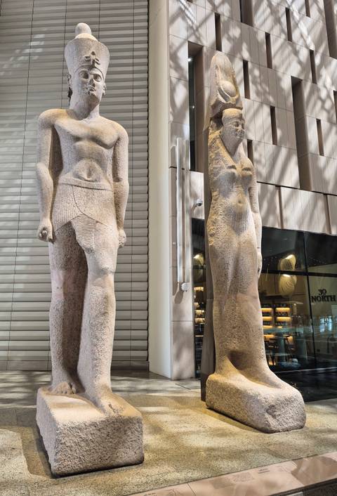 Two colossal ancient statues displayed upright inside a brightly lit modern museum atrium.