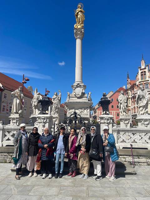 Tour group poses in front of ornate baroque column surrounded by stone statues in a European square