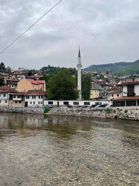 Riverbank view of Bosnian town with hillside houses and slender minaret under grey skies