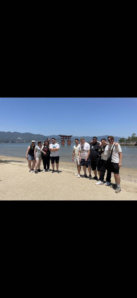 Tour group poses on a sandy shore with the famous floating torii gate of Miyajima island in the background.