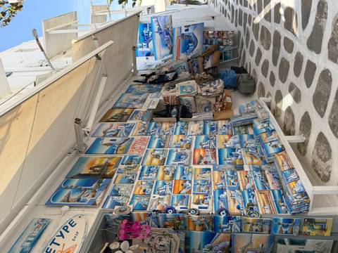 Outdoor stall displaying dozens of colorful Greek landscape paintings and souvenirs along a white stone path.