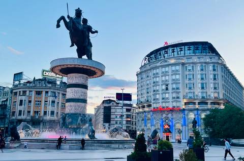 An illuminated fountain surrounds a heroic equestrian statue in a lively city square at dusk.