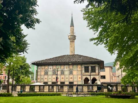 A richly painted Ottoman-style mosque with a tall slender minaret framed by leafy trees on a grey day.