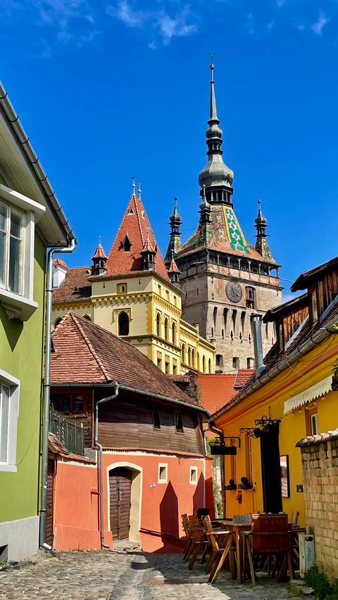 Turreted medieval buildings and a colourful clock tower rise above tiled rooftops under a clear blue sky.